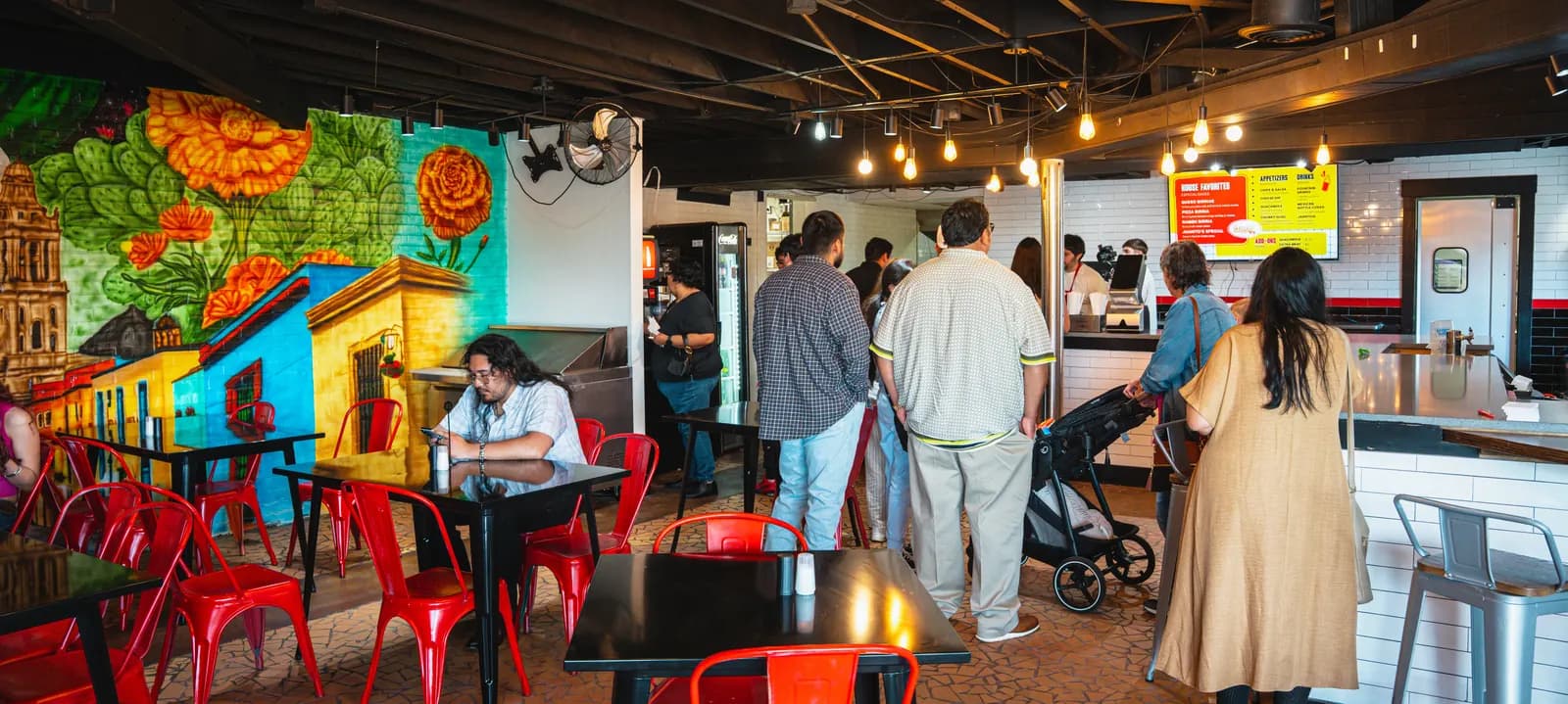 The dining room at El Callejón GTO: mural-painted walls, red metal chairs and a line of guests ordering at the counter under the menu board